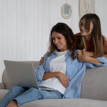 Happy smiling family of woman and teen girl watching interesting video in laptop in living room